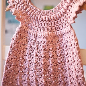 Peut inclure: Une robe de b&eacute;b&eacute; en crochet rose avec un ourlet festonn&eacute; et des manches courtes. La robe pr&eacute;sente un motif floral d&eacute;licat et est r&eacute;alis&eacute;e avec un fil doux.