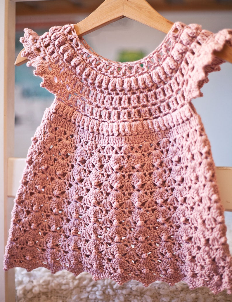Peut inclure: Une robe de b&eacute;b&eacute; en crochet rose avec un ourlet festonn&eacute; et des manches courtes. La robe pr&eacute;sente un motif floral d&eacute;licat et est r&eacute;alis&eacute;e avec un fil doux.