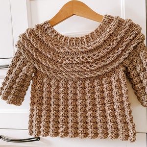 K&ouml;nnte beinhalten: Ein brauner H&auml;kelpullover mit einem breiten, gekr&auml;uselten Ausschnitt und langen &Auml;rmeln. Der Pullover ist mit einem strukturierten Stich gearbeitet, der eine erhabene, unebene Oberfl&auml;che erzeugt.