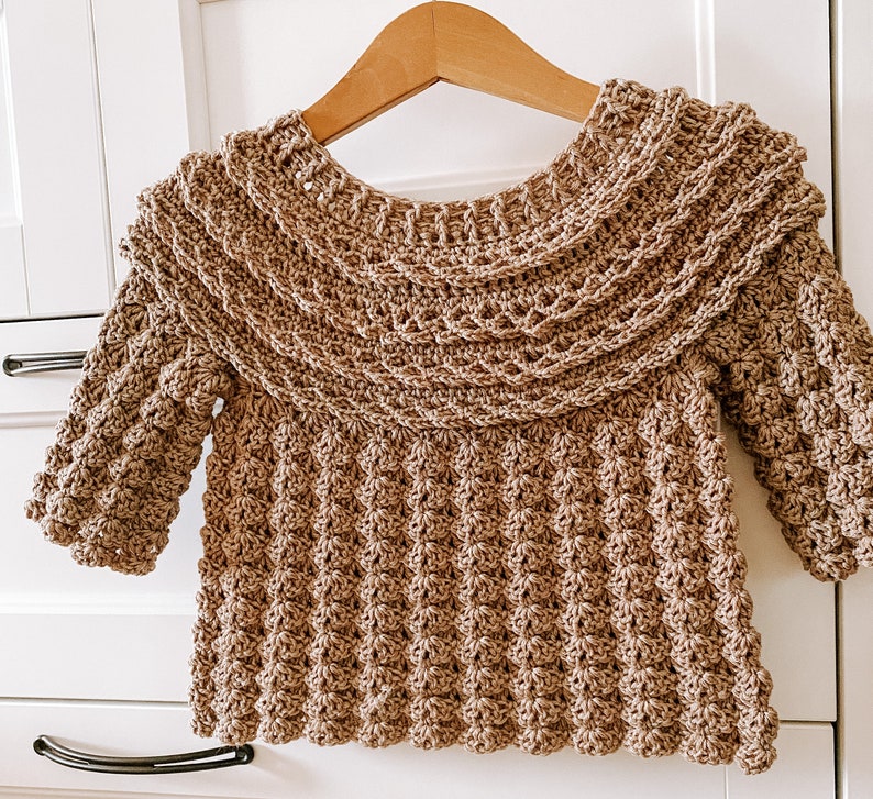 K&ouml;nnte beinhalten: Ein brauner H&auml;kelpullover mit einem breiten, gekr&auml;uselten Ausschnitt und langen &Auml;rmeln. Der Pullover ist mit einem strukturierten Stich gearbeitet, der eine erhabene, unebene Oberfl&auml;che erzeugt.