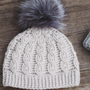 May include: A cream-colored knitted beanie with a cable knit pattern and a fluffy gray pom-pom on top.