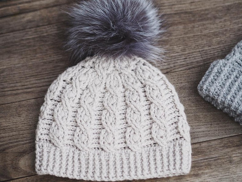 May include: A cream-colored knitted beanie with a cable knit pattern and a fluffy gray pom-pom on top.
