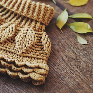 May include: A close-up of a crocheted garment with a textured, leaf-like pattern in a warm brown color. The garment is lying on a wooden surface.