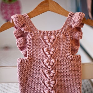 May include: A pink crocheted baby top with ruffled sleeves and a textured design featuring a series of small, raised balls.