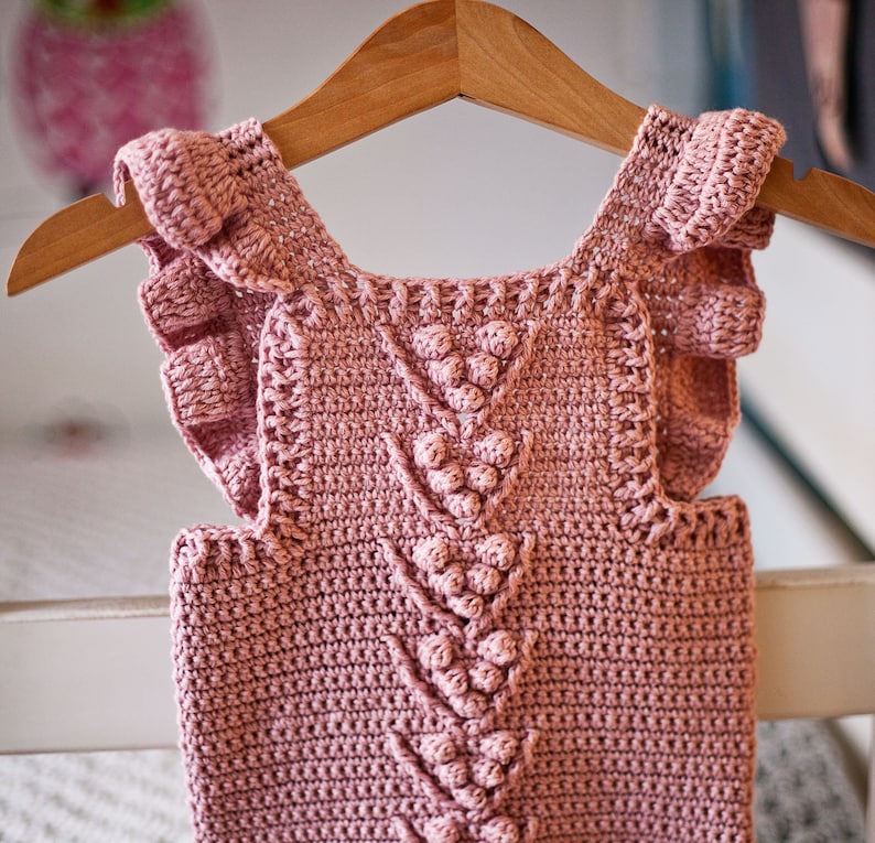 May include: A pink crocheted baby top with ruffled sleeves and a textured design featuring a series of small, raised balls.