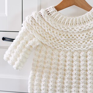 K&ouml;nnte beinhalten: Ein wei&szlig;er H&auml;kelpullover mit einem R&uuml;schenkragen und einem strukturierten Design. Der Pullover h&auml;ngt an einem Holzb&uuml;gel.