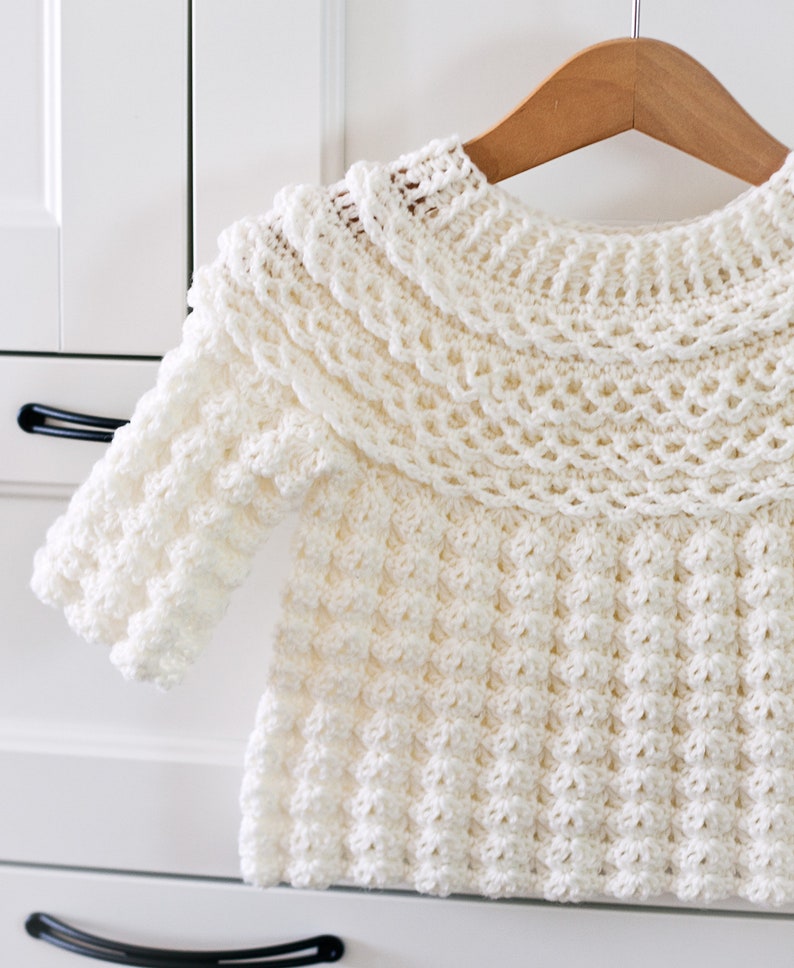 K&ouml;nnte beinhalten: Ein wei&szlig;er H&auml;kelpullover mit einem R&uuml;schenkragen und einem strukturierten Design. Der Pullover h&auml;ngt an einem Holzb&uuml;gel.