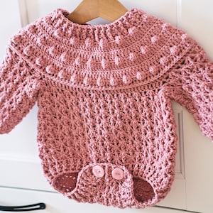 May include: A pink crocheted baby onesie with a ruffled neckline and a textured design. The onesie has a button closure at the bottom.