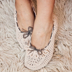 Può includere: Un paio di pantofole all'uncinetto di colore beige chiaro con dettagli in nastro grigio. Le pantofole sono indossate su un tappeto bianco e soffice.