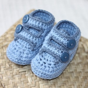 Puede incluir: Un par de botitas de bebé de crochet azul con botones azules. Las botitas tienen una correa en la parte superior y están hechas con un punto texturizado.