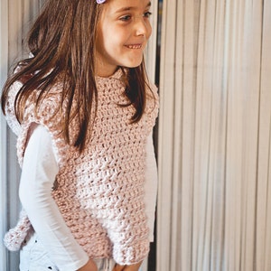 K&ouml;nnte beinhalten: Ein junges M&auml;dchen tr&auml;gt eine rosa geh&auml;kelte Weste mit einem wei&szlig;en Langarmshirt darunter. Sie steht vor einem wei&szlig;en Vorhang.