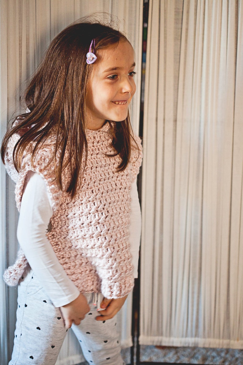 K&ouml;nnte beinhalten: Ein junges M&auml;dchen tr&auml;gt eine rosa geh&auml;kelte Weste mit einem wei&szlig;en Langarmshirt darunter. Sie steht vor einem wei&szlig;en Vorhang.