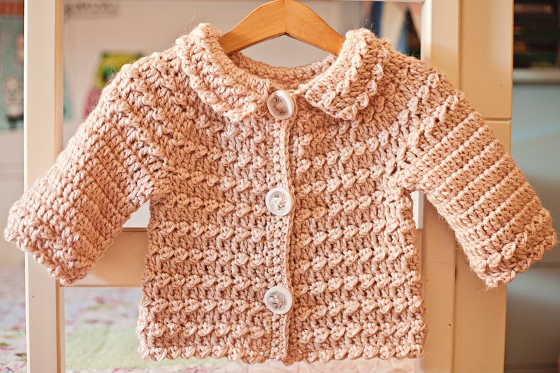 May include: A light pink, crocheted baby cardigan with a scalloped collar and three white buttons. The long-sleeved sweater is displayed on a wooden hanger, showcasing the textured stitch pattern.