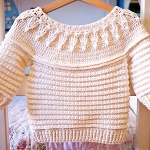 May include: Cream-colored crochet sweater with long sleeves, hanging on a wooden hanger. The sweater features a wide, decorative collar and ribbed detailing on the sleeves and body. The handmade garment is displayed against a soft, floral background.