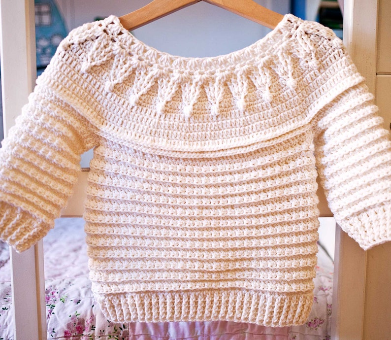 May include: Cream-colored crochet sweater with long sleeves, hanging on a wooden hanger. The sweater features a wide, decorative collar and ribbed detailing on the sleeves and body. The handmade garment is displayed against a soft, floral background.