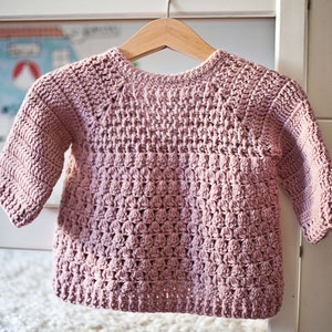 May include: A pink crocheted sweater with a textured stitch pattern. The sweater has long sleeves and a round neckline.