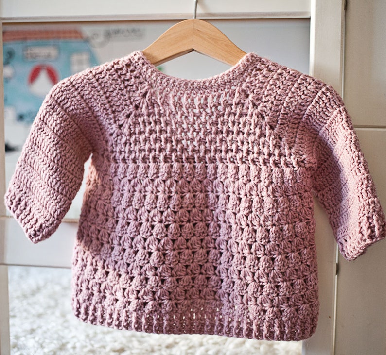 May include: A pink crocheted sweater with a textured stitch pattern. The sweater has long sleeves and a round neckline.
