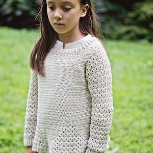 K&ouml;nnte beinhalten: Ein hellbeige H&auml;kelpullover mit langen &Auml;rmeln. Der Pullover hat ein strukturiertes offenes Webmuster und eine leicht ausgestellte Form. Das Model tr&auml;gt ein wei&szlig;es Hemd darunter und ein blaues Blumenstirnband. Der Hintergrund ist verschwommenes gr&uuml;nes Gras.