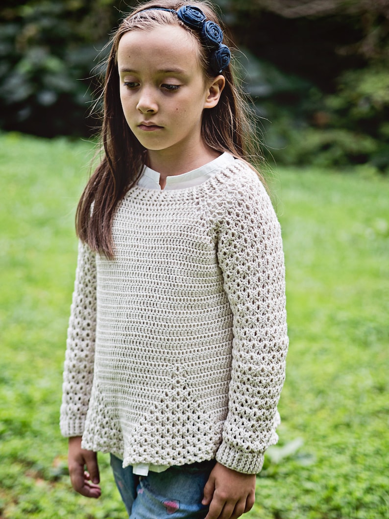 K&ouml;nnte beinhalten: Ein hellbeige H&auml;kelpullover mit langen &Auml;rmeln. Der Pullover hat ein strukturiertes offenes Webmuster und eine leicht ausgestellte Form. Das Model tr&auml;gt ein wei&szlig;es Hemd darunter und ein blaues Blumenstirnband. Der Hintergrund ist verschwommenes gr&uuml;nes Gras.