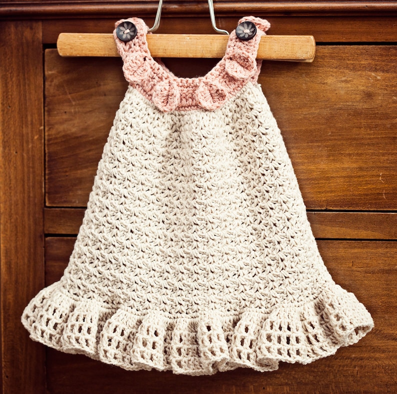 Op de afbeelding: Een gehaakt babyjurkje met een roze gerimpelde halslijn en een beige gehaakte rok. Het jurkje hangt aan een houten hanger.