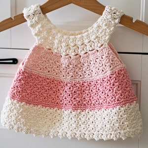 May include: A crocheted baby dress with a white bodice and pink skirt. The dress has a scalloped edge and is made with a delicate stitch.