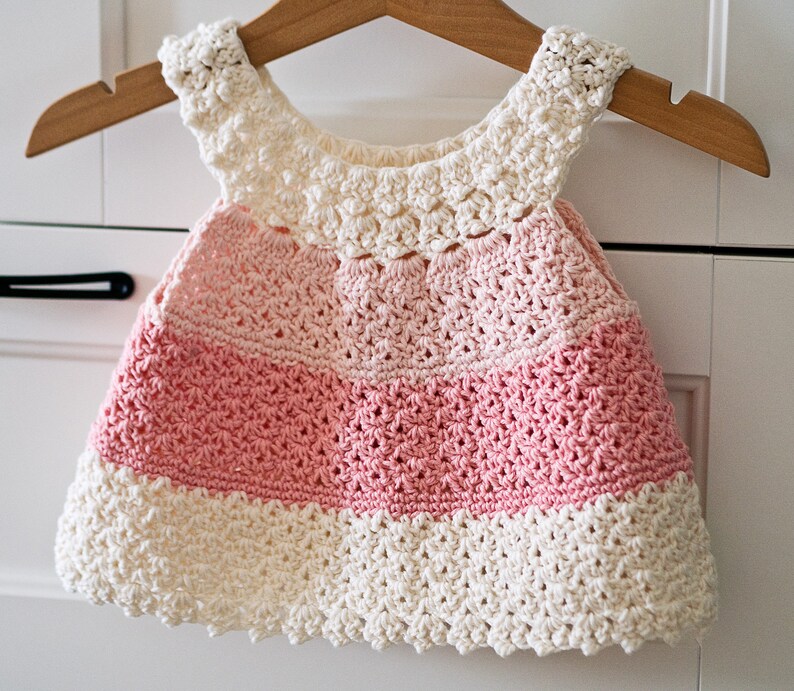 May include: A crocheted baby dress with a white bodice and pink skirt. The dress has a scalloped edge and is made with a delicate stitch.