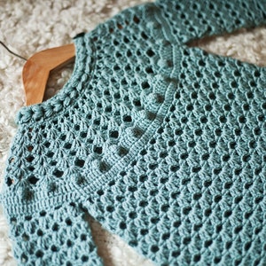 Puede incluir: Un su&eacute;ter de crochet azul claro con un delicado patr&oacute;n de calado. El su&eacute;ter tiene un cuello redondo y mangas largas.