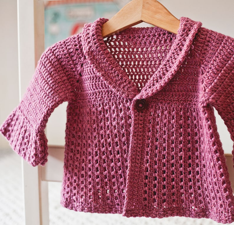 May include: A pink crocheted cardigan with a button closure and a wide collar. The cardigan has a lacy pattern and is made with a soft yarn.