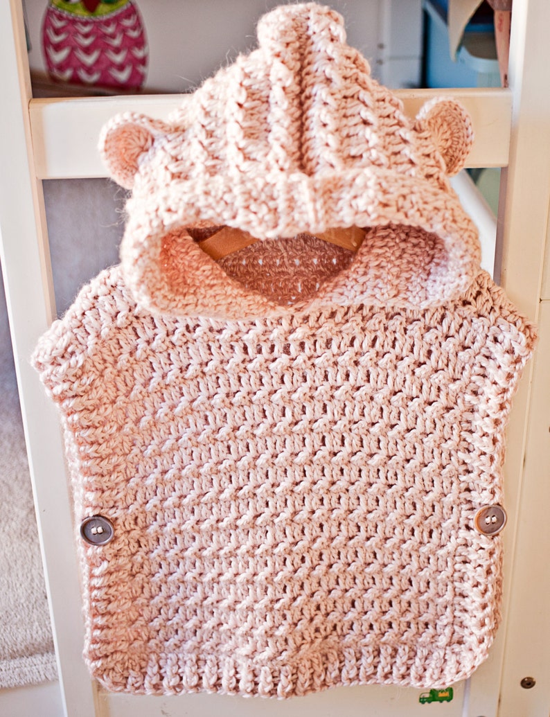 K&ouml;nnte beinhalten: Eine rosa geh&auml;kelte Kapuzenweste mit B&auml;renohren und Kn&ouml;pfen. Die Weste ist mit einem strukturierten Stichmuster gearbeitet.