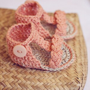 Puede incluir: Un par de sandalias de beb&eacute; de crochet rosa con un detalle de bot&oacute;n. Las sandalias est&aacute;n sobre una cesta tejida.