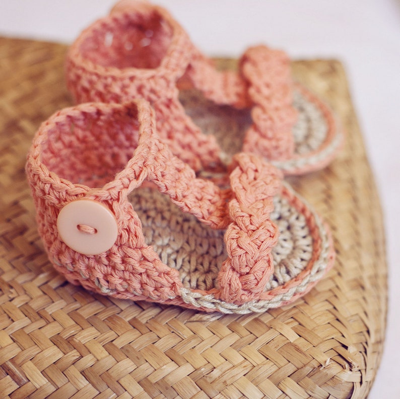 Puede incluir: Un par de sandalias de beb&eacute; de crochet rosa con un detalle de bot&oacute;n. Las sandalias est&aacute;n sobre una cesta tejida.