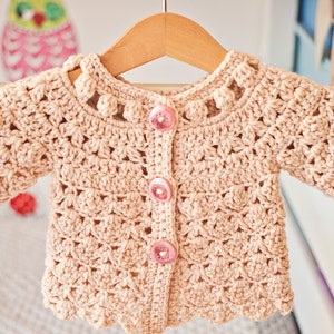 May include: A crocheted baby cardigan in a light beige color with pink buttons and a scalloped hem. The cardigan has a delicate crochet pattern and is hung on a wooden hanger.