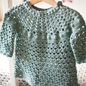 Puede incluir: Un su&eacute;ter de crochet azul claro con un delicado patr&oacute;n de calado. El su&eacute;ter tiene un cuello redondo y mangas tres cuartos.