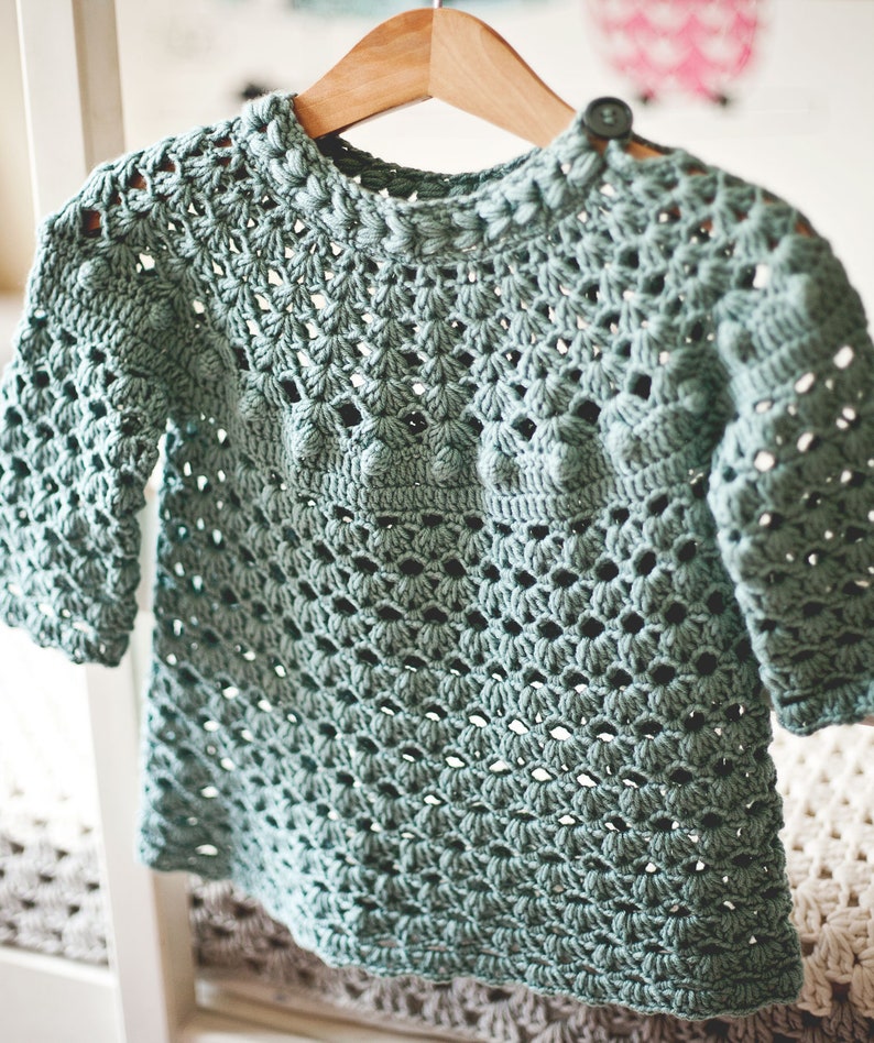 Puede incluir: Un su&eacute;ter de crochet azul claro con un delicado patr&oacute;n de calado. El su&eacute;ter tiene un cuello redondo y mangas tres cuartos.