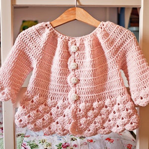 May include: A pink crocheted cardigan with a scalloped hem and white buttons. The cardigan has long sleeves and a round neckline.