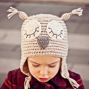 May include: A child wearing a beige crocheted owl hat with brown eyes and a gray nose. The hat has ear flaps and ties that hang down the back of the neck.