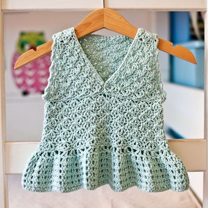 May include: A light green crocheted sleeveless top with a ruffled hem. The top is hanging on a wooden hanger.