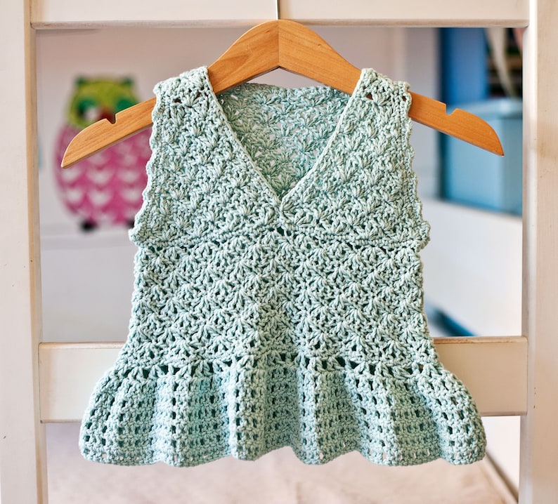 May include: A light green crocheted sleeveless top with a ruffled hem. The top is hanging on a wooden hanger.