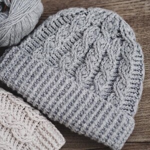 May include: Two crocheted hats, one gray and one beige, with a cable knit pattern. The hats are lying on a wooden surface.