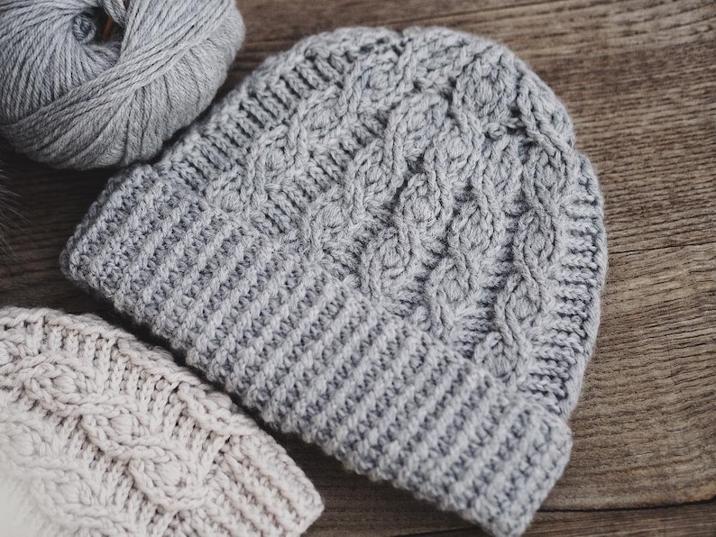 May include: Two crocheted hats, one gray and one beige, with a cable knit pattern. The hats are lying on a wooden surface.