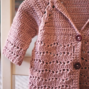 May include: A dusty rose crocheted cardigan with a button closure. The cardigan has a delicate open stitch pattern and is hanging on a wooden hanger.