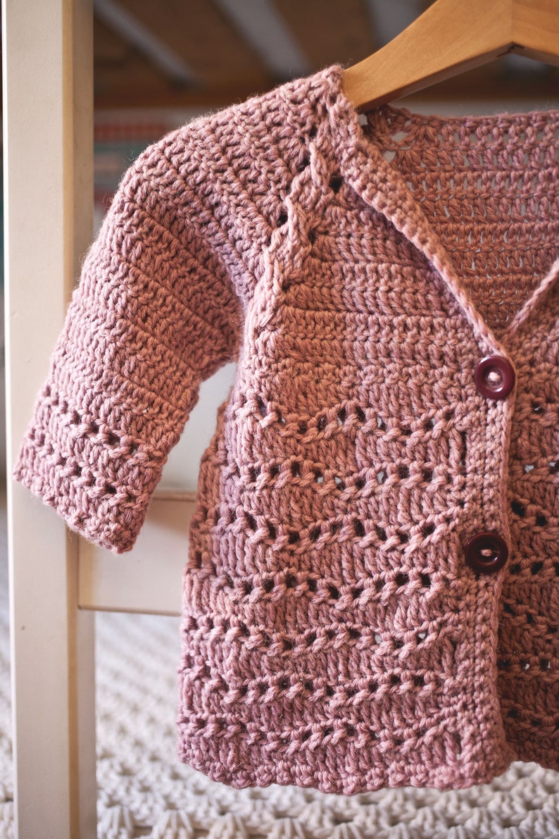 May include: A dusty rose crocheted cardigan with a button closure. The cardigan has a delicate open stitch pattern and is hanging on a wooden hanger.