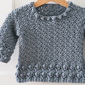 May include: A light blue crocheted sweater with a textured stitch pattern and a row of crocheted bobbles around the neckline and hem.