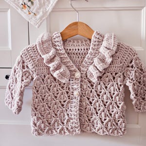 May include: A crocheted baby cardigan in a pale pink colour with a ruffled collar and button fastening.