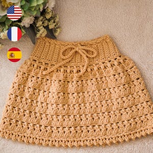 May include: A mustard yellow crochet skirt featuring a ribbed waistband with a matching drawstring tie. The skirt displays a repeating textured pattern of vertical stitches and small bobble clusters, finished with a scalloped bottom edge.