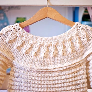 May include: Cream-colored, handmade crochet sweater with long sleeves, hanging on a wooden hanger. The sweater features a detailed, textured stitch pattern, with a decorative collar and a scalloped edge. The garment is likely for a baby or toddler.