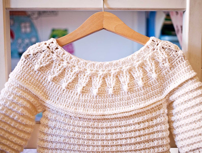 May include: Cream-colored, handmade crochet sweater with long sleeves, hanging on a wooden hanger. The sweater features a detailed, textured stitch pattern, with a decorative collar and a scalloped edge. The garment is likely for a baby or toddler.