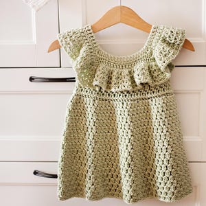 Può includere: Un abito in crochet verde chiaro con scollo e maniche a balze. L'abito è appeso a un appendiabiti in legno.