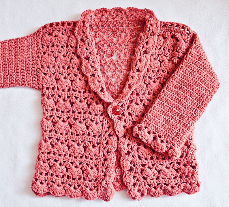 May include: A coral-colored, crocheted cardigan with a scalloped collar and hem. The long-sleeved garment features a single red button closure. The openwork design is visible throughout the sweater, creating a delicate, textured appearance.