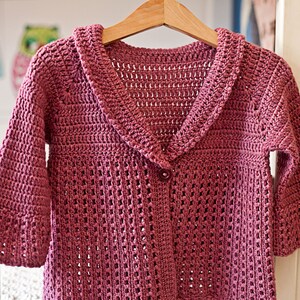 May include: A pink crocheted cardigan with a wide collar and button closure. The cardigan has a delicate openwork pattern.
